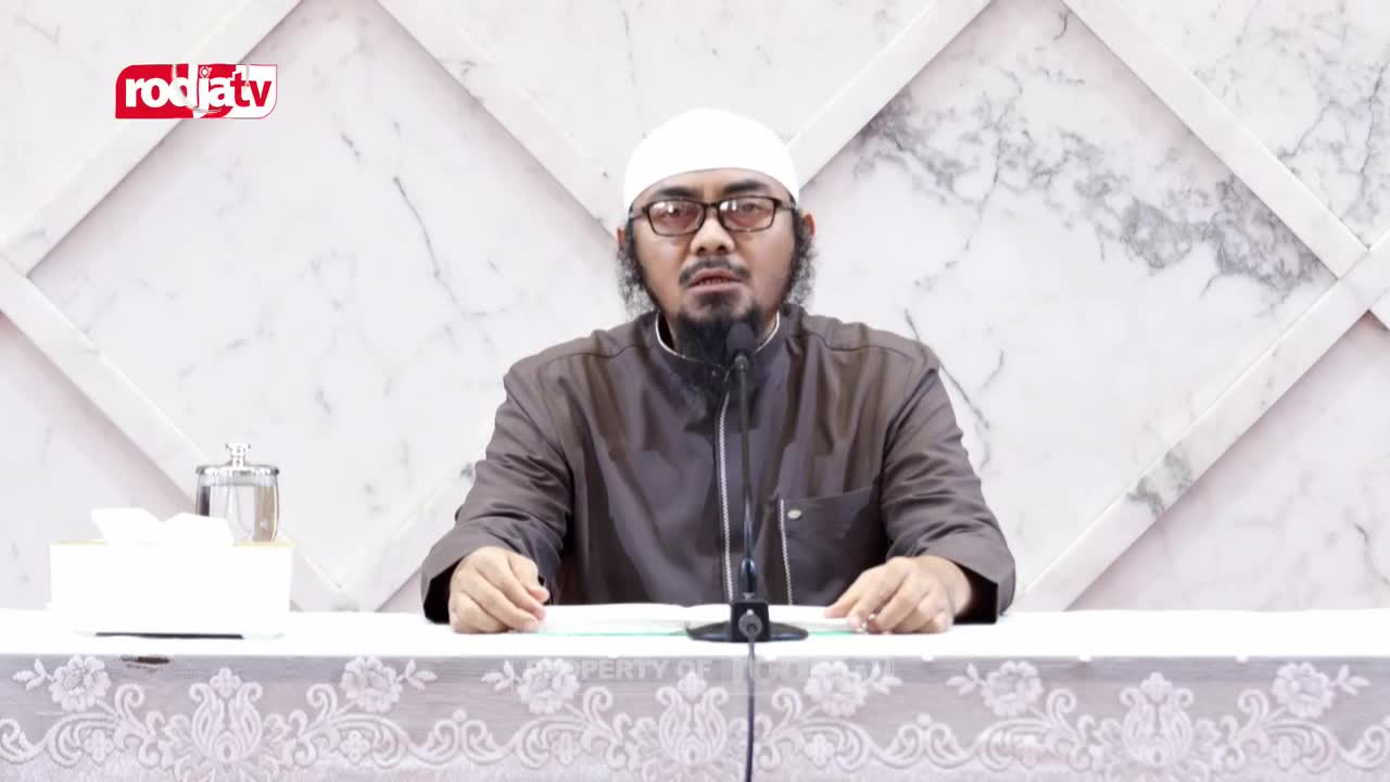 A man in a white cap and glasses speaks into a microphone, his hands resting on a book. The Rodja TV logo is visible in the corner, and the backdrop is a marble-patterned wall.
