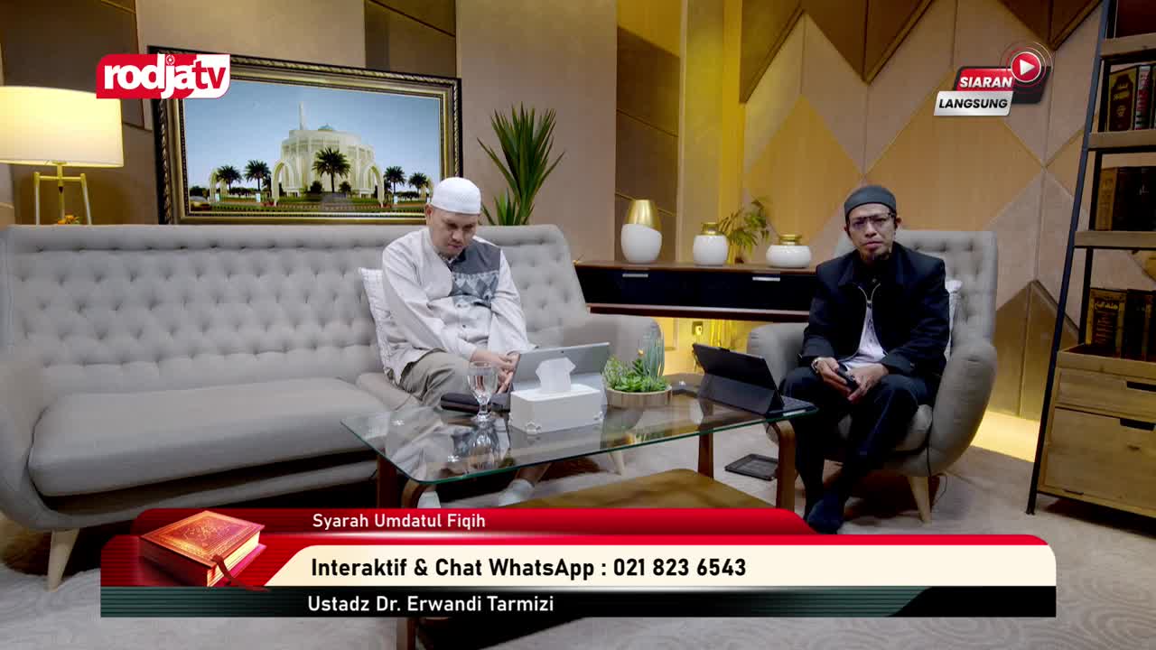 Two men sit on a set, one on a sofa and the other in a chair, both facing forward. A Rodja TV logo and "Siaran Langsung" are visible, with a lower third banner advertising a live program from Indonesia.

