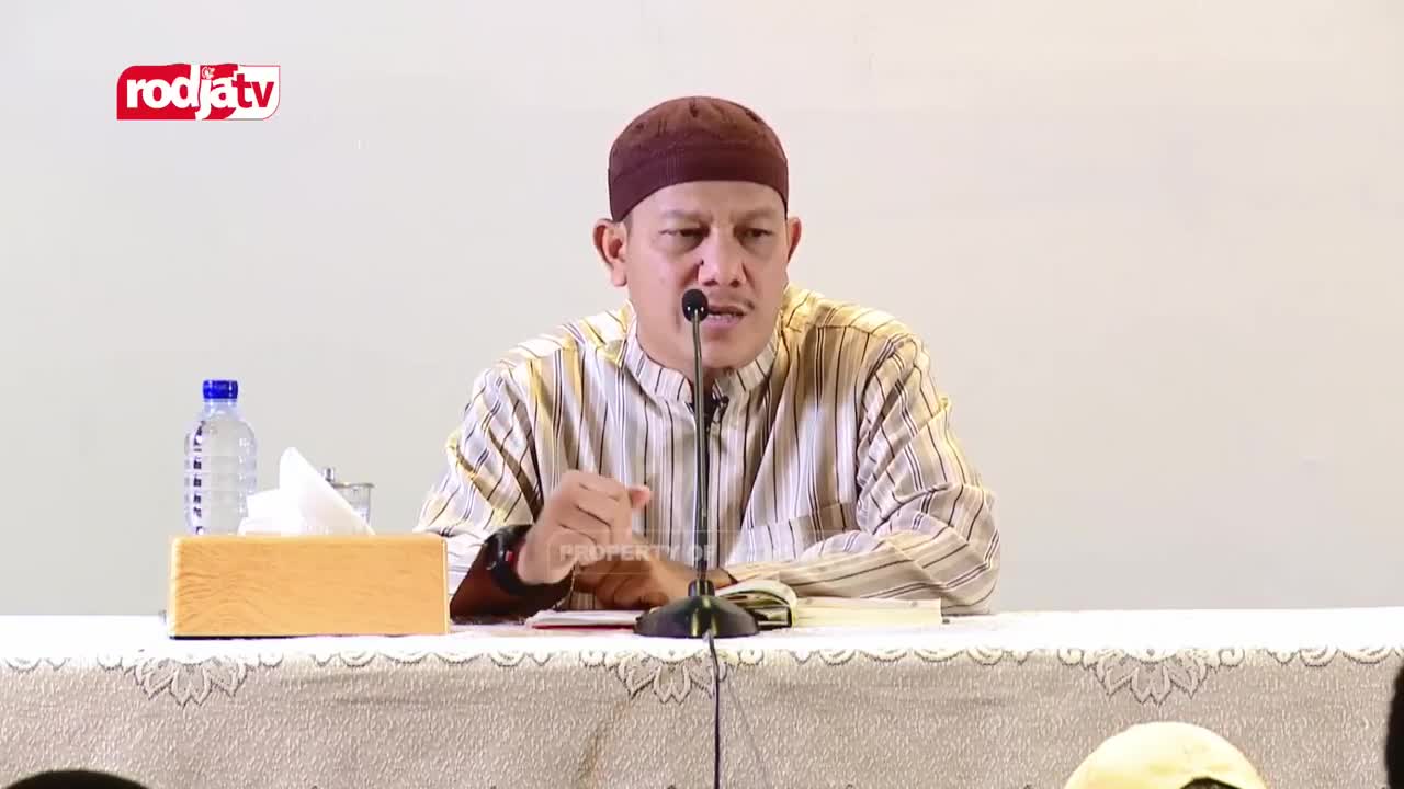 A man in a striped shirt and cap speaks into a microphone, gesturing with his hands.  The Rodja TV logo is visible in the corner, and a bottle of water and tissues sit on the table.
