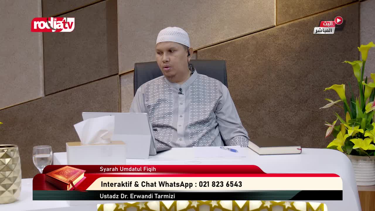 A man in a white cap and grey shirt is speaking, seated at a desk on what appears to be a Rodja TV broadcast. A graphic at the bottom identifies the program and how to contact them.
