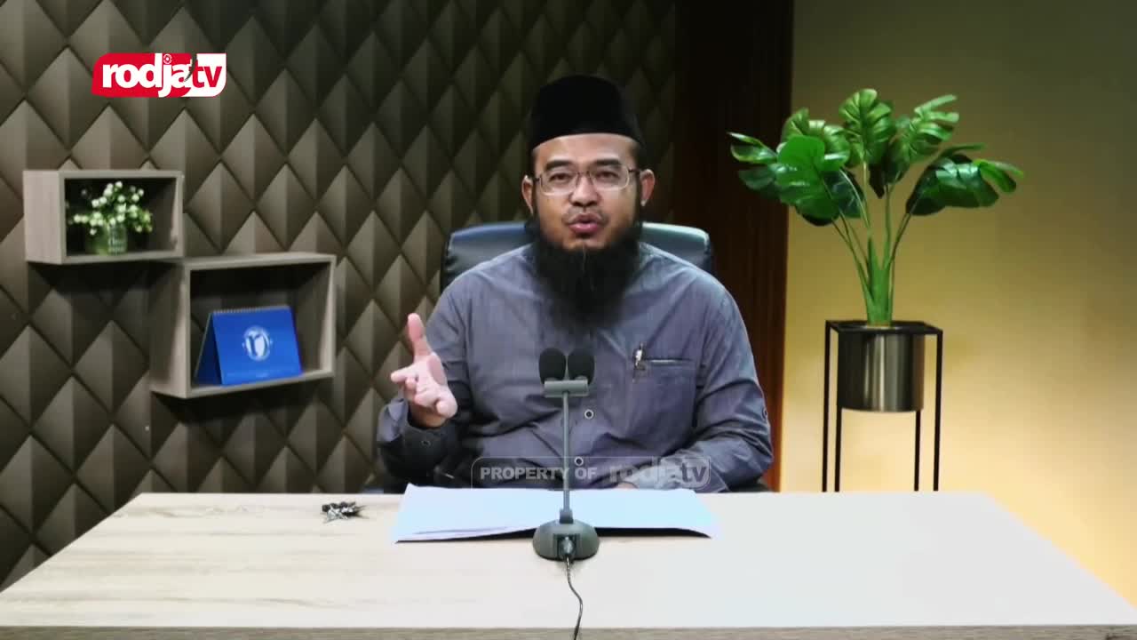 A man in a black cap and glasses gestures with his right hand, speaking into two microphones. The Rodja TV logo is visible in the corner, and the studio backdrop is a textured wall.
