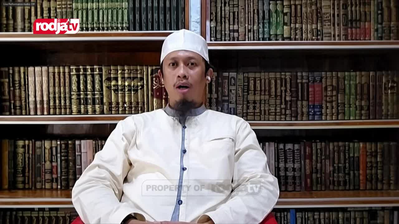 A man in a white peci and traditional Indonesian clothing speaks directly to the camera, his mouth slightly open. Behind him, shelves packed with books stretch across the background, a familiar sight on Rodja TV.