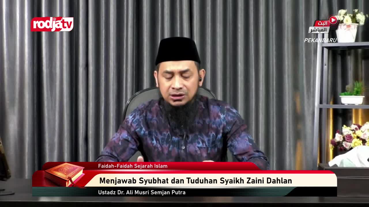 A man wearing a peci and a patterned shirt speaks from behind a desk. A Rodja TV logo appears in the upper left corner of the screen. A man wearing a peci and a patterned shirt speaks from behind a desk. A Rodja TV logo appears in the upper left corner of the screen.