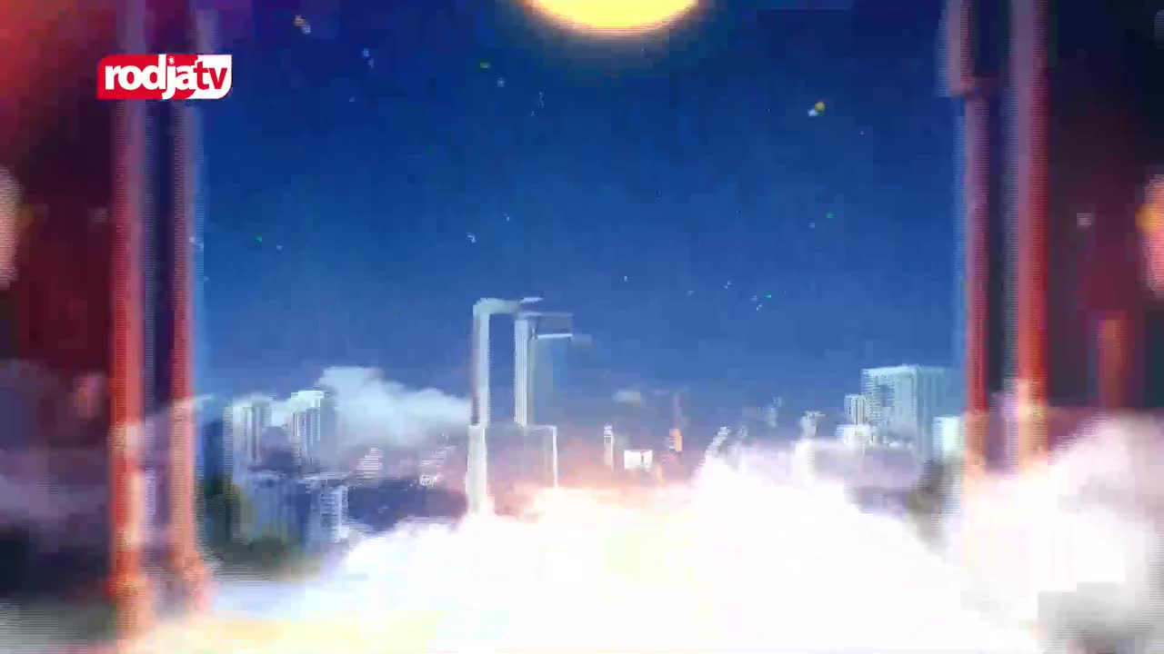 A vast, bright sun hangs in the deep blue sky above a hazy cityscape. The Rodja TV logo is visible in the upper left corner. A vast, bright sun hangs in the deep blue sky above a hazy cityscape. The Rodja TV logo is visible in the upper left corner.