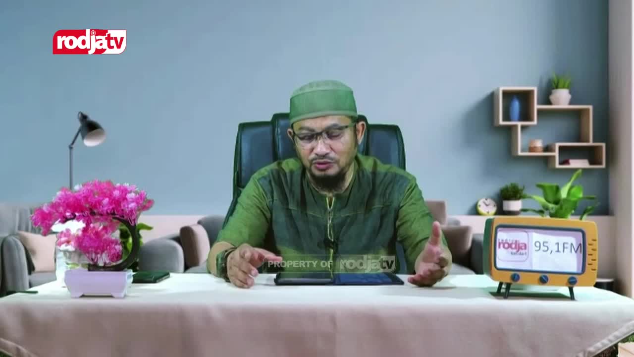 A man in a green cap and shirt gestures with his hands while speaking from behind a desk. A vintage-style radio displaying "Rodja 95,1 FM" sits on the table in front of him. A man in a green cap and shirt gestures with his hands while speaking from behind a desk. A vintage-style radio displaying "Rodja 95,1 FM" sits on the table in front of him.