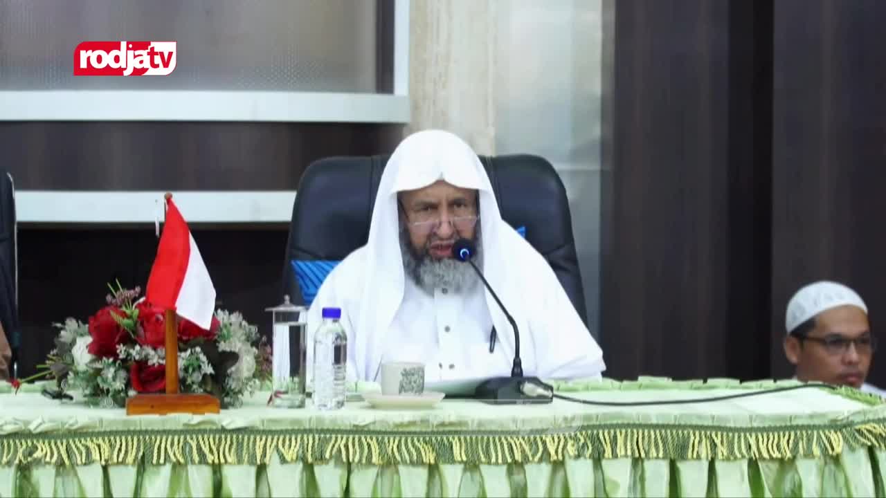 A man in a white robe and head covering speaks into a microphone, his gaze directed forward. Beside him, the Indonesian flag stands proudly amongst a bouquet of red and white flowers. A man in a white robe and head covering speaks into a microphone, his gaze directed forward. Beside him, the Indonesian flag stands proudly amongst a bouquet of red and white flowers.