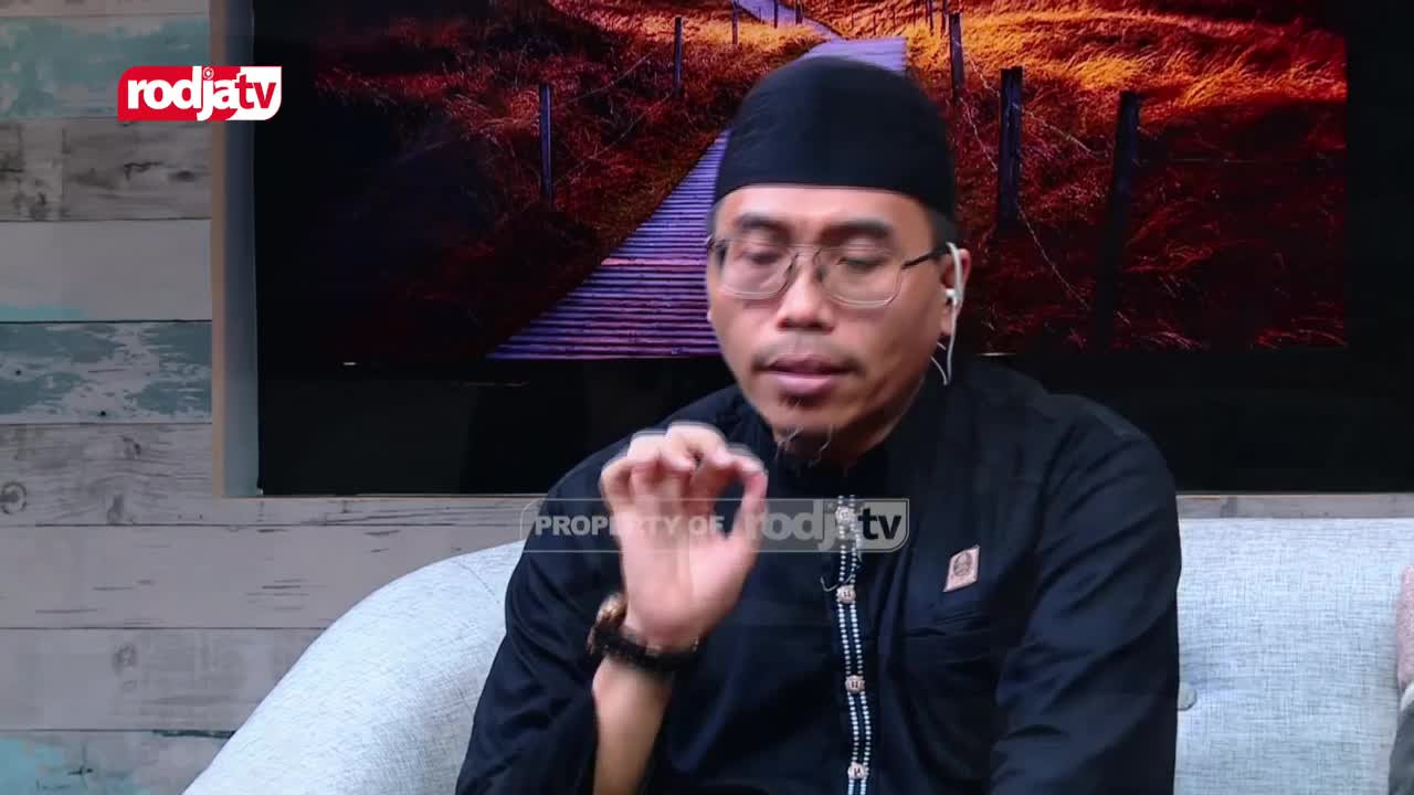 A man in a black peci and glasses speaks, gesturing with his right hand. Behind him, a Rodja TV logo appears on a screen showing a pathway through dry grass.