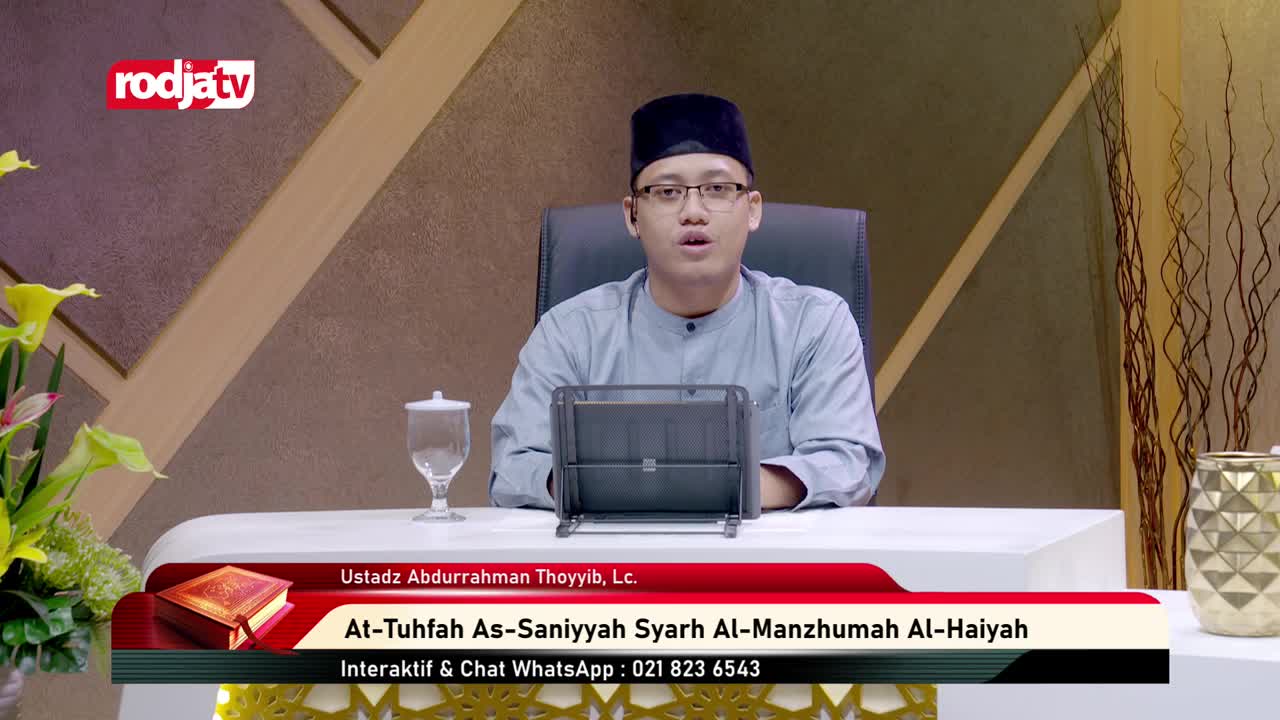 A man in a peci and glasses speaks from behind a small tablet, his voice broadcast by Rodja TV. A glass of water sits to his left, and a decorative vase to his right.