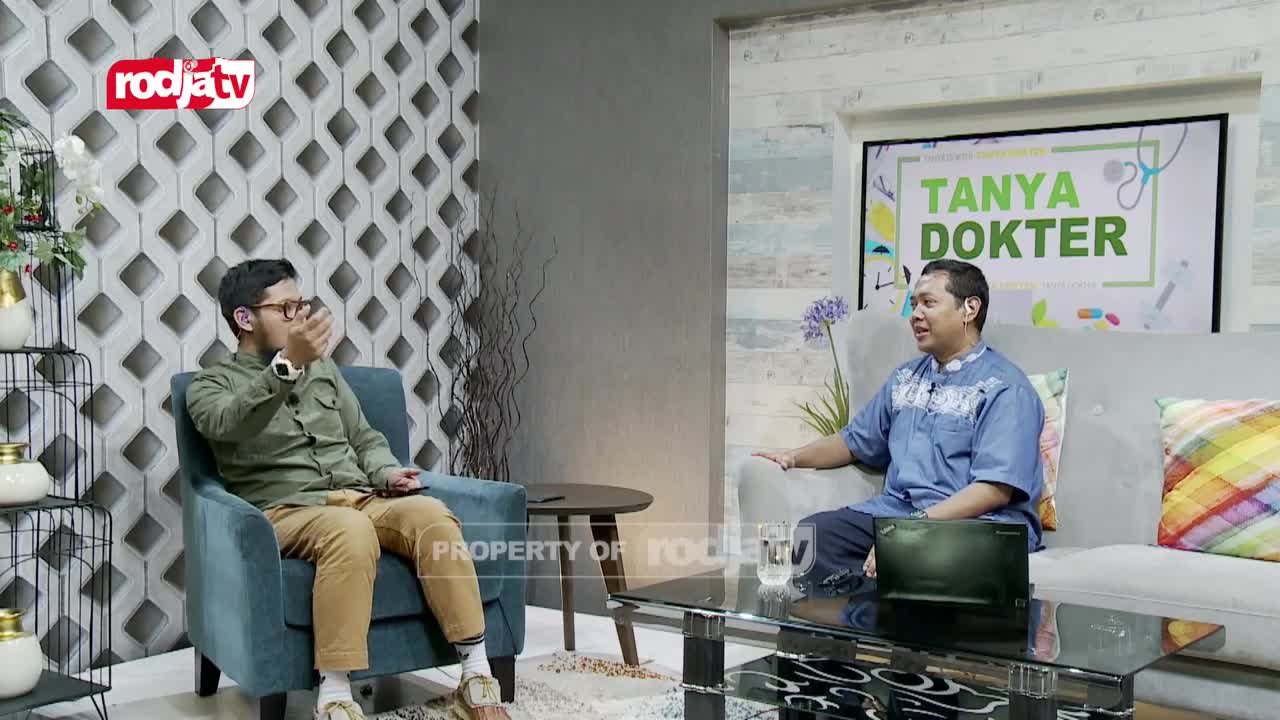 A man in a green shirt gestures while speaking, seated in a teal armchair. Across from him, a man in a blue tunic smiles, leaning back on a gray sofa, with a laptop on the coffee table between them. A screen behind them displays "TANYA DOKTER," a program from Rodja TV.