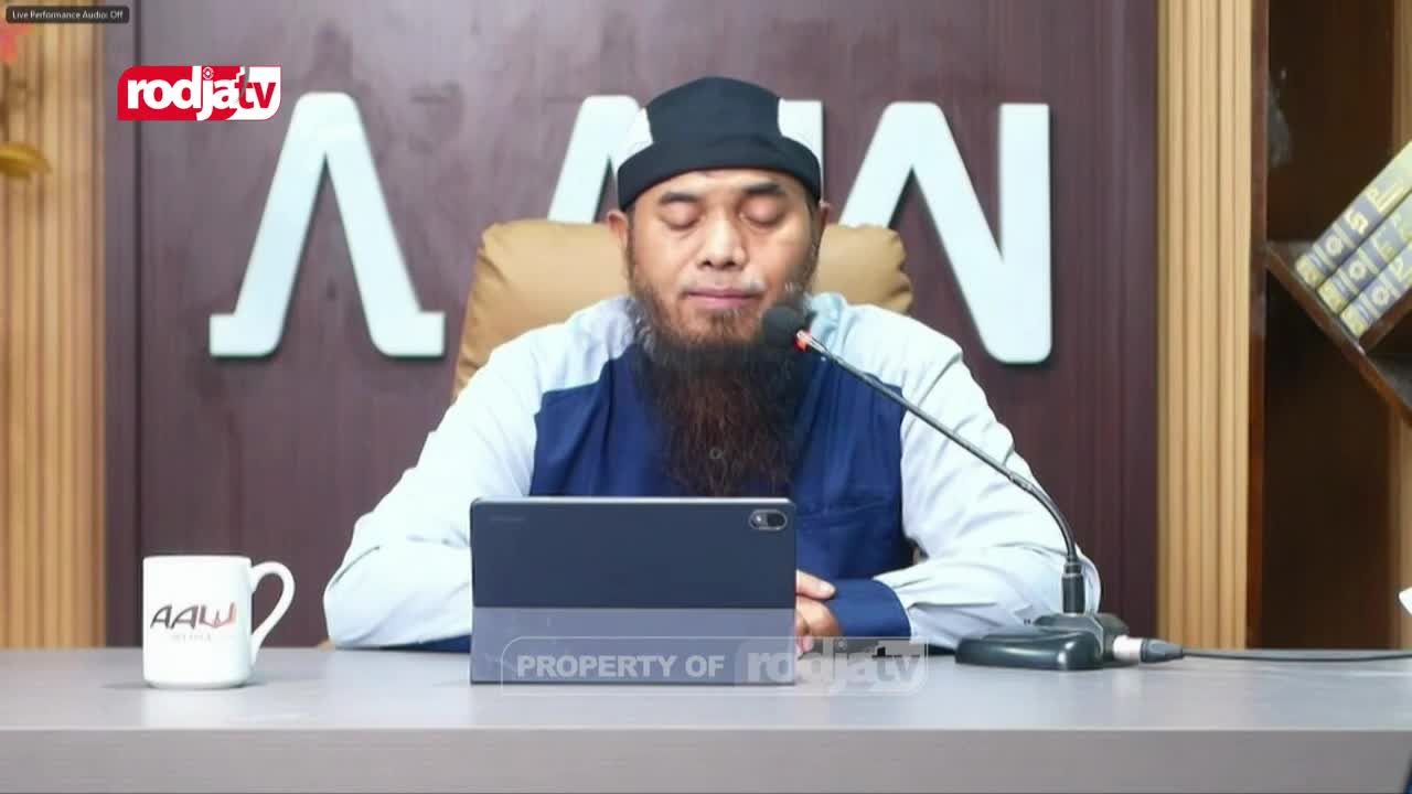 A man with a long beard, wearing a black cap and a blue and white shirt, speaks into a microphone. He sits behind a table with a tablet and a mug bearing the "AAW" logo.