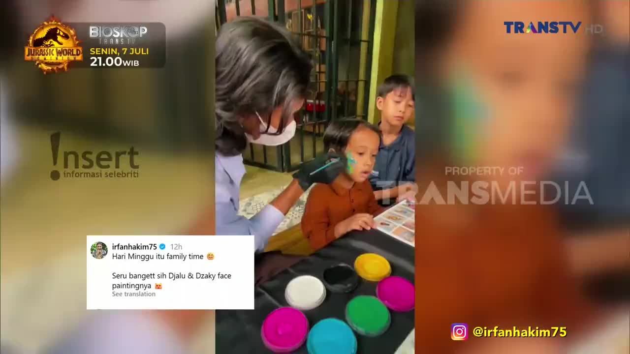 A person wearing a mask is applying green face paint to a child's cheek. Another child watches as colorful paints sit on a table, and the Trans TV logo is visible in the background.
