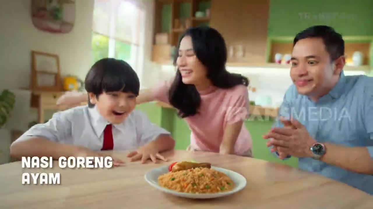 A family is gathered around a table, smiling at a plate of nasi goreng ayam. The scene, likely from a commercial on Trans TV in Indonesia, exudes warmth and joy.
