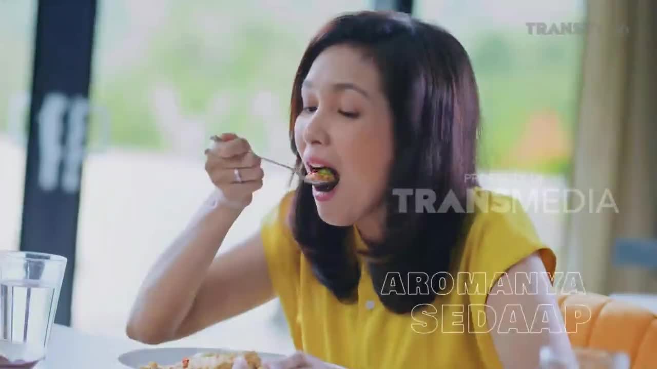 A woman in a yellow top is mid-bite, a forkful of food heading toward her open mouth. The scene, with the Trans TV logo visible, appears to be from a cooking show in Indonesia.
