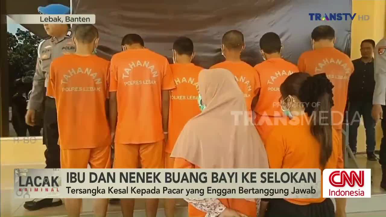 Eight men in matching orange jumpsuits stand in a line, their backs to the camera. A woman in a hijab and another with a face mask are positioned in front of them, with the Trans TV logo visible in the background.
