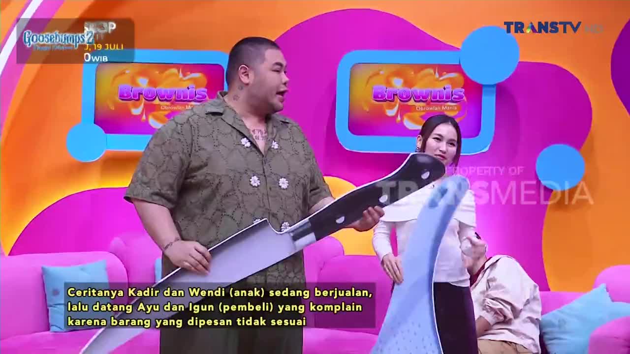 A man on the Indonesian Trans TV show "Brownis" holds a comically oversized knife, gesturing with it. A woman stands beside him, holding a blue object, while another person is partially visible in the foreground.
