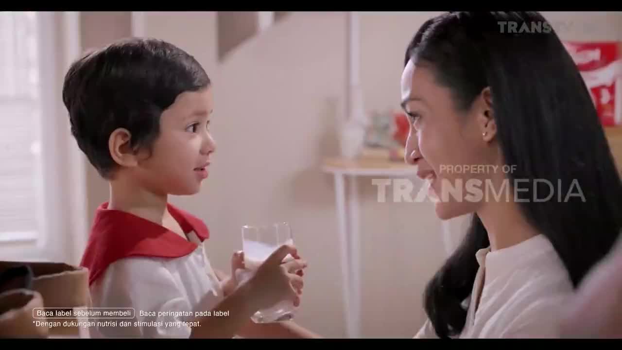 A young child, holding a glass of milk, looks up at a woman who is smiling. The scene appears to be part of a commercial on Trans TV.
