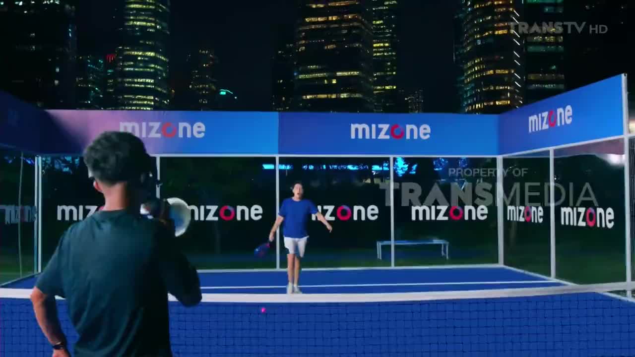 A player in a blue shirt prepares to hit a pink ball on a blue court, with "mizone" branding visible. The scene is set at night, with a camera operator capturing the action for Trans TV Indonesia.
