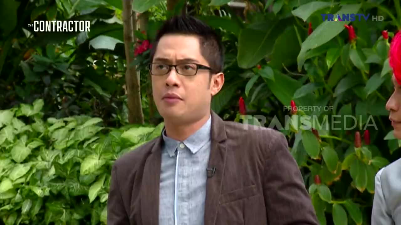 A man in glasses and a blazer is looking intently at something off-camera, his expression serious. The scene appears to be a segment from "The Contractor" on Trans TV, with lush greenery in the background.
