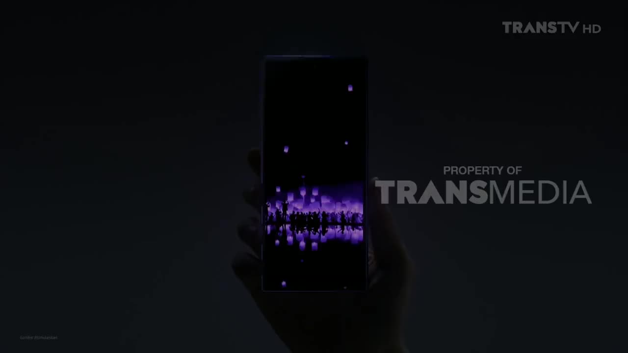 A hand holds a phone displaying a purple scene of floating lanterns. The Trans TV logo and Transmedia property notice are visible.
