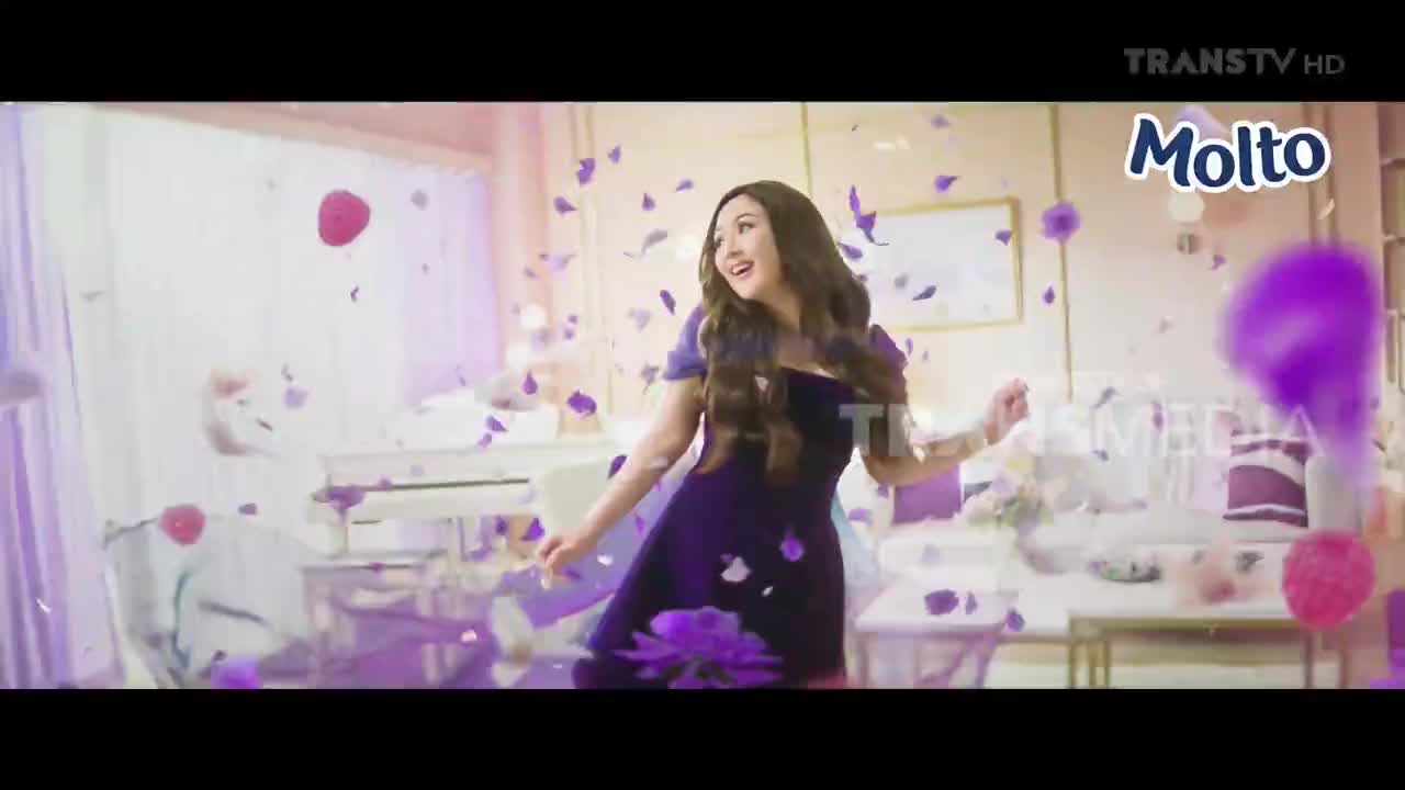 A woman in a dark purple dress dances in a room as flower petals float around her. The scene is part of a commercial on Trans TV in Indonesia, promoting Molto.
