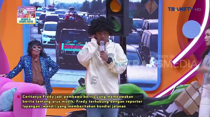 On a Trans TV set, a man with a microphone and a large afro is holding his earpiece, likely listening to someone. Behind him, a backdrop shows a traffic jam, and a man in a sparkly jacket leans on a pink couch.
