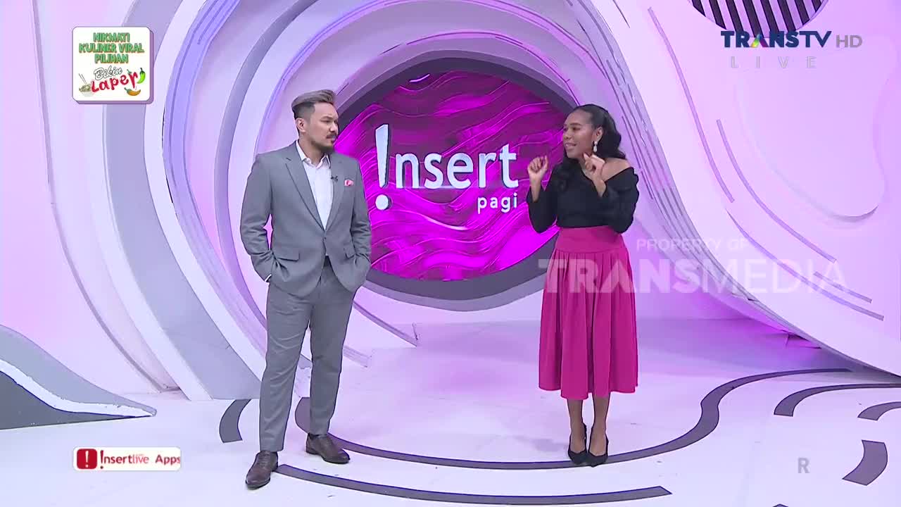 The woman in the pink skirt gestures emphatically while talking, her co-host stands calmly beside her. The set is brightly lit, with the "Insert Pagi" logo clearly visible on the screen from Trans TV.
