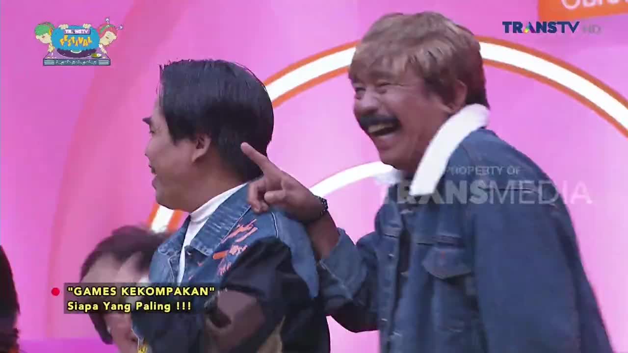 On the Trans TV set, a man in a denim jacket points and laughs, his face crinkling with amusement. Beside him, another man in a similar jacket looks on, seemingly the target of the joke.
