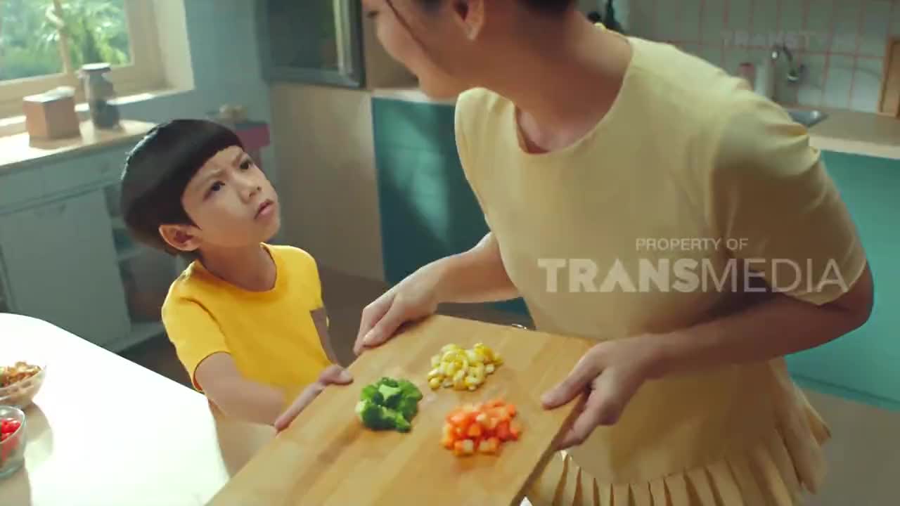 A woman holds a wooden board with chopped vegetables, showing them to a boy in a yellow shirt. The boy looks up at her with a thoughtful expression.
