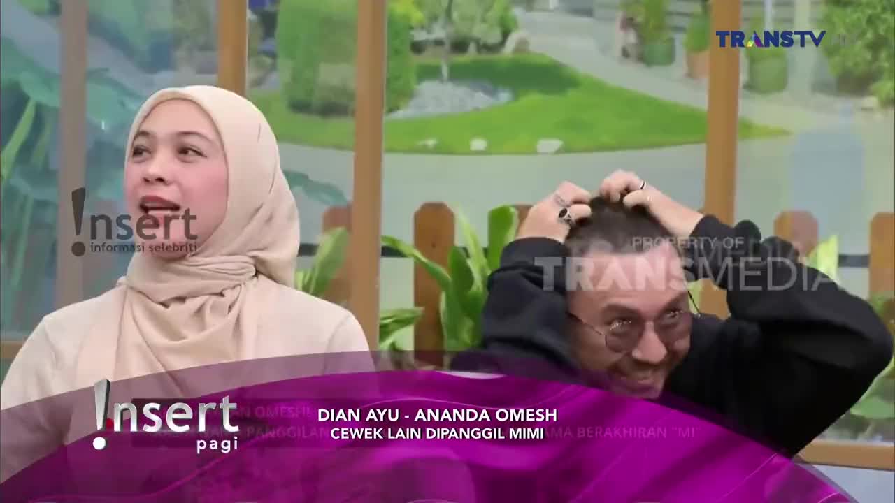 On the Trans TV set, the woman in the hijab is speaking animatedly. Across from her, a man with glasses scratches his head, looking amused.
