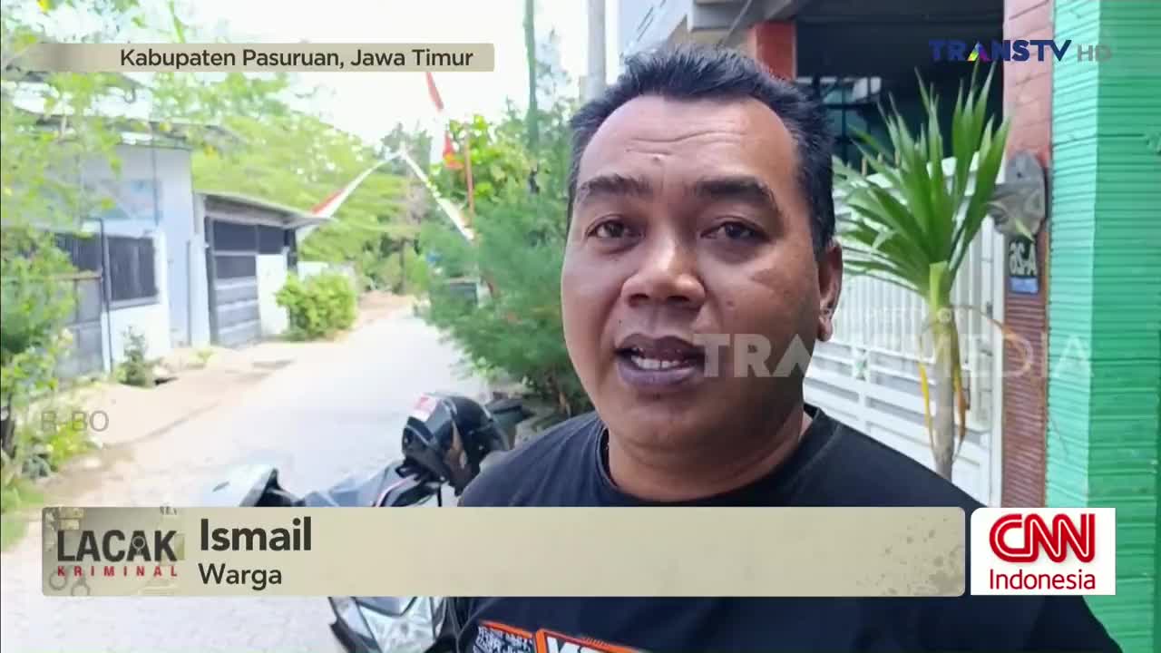 A man is speaking directly to the camera, with a Trans TV logo visible in the corner. Behind him, a motorcycle is parked on a quiet street in Kabupaten Pasuruan, Java Timur.
