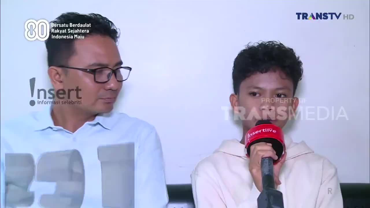 A young man holds a red microphone, speaking into it, while another man in glasses looks on. The scene is likely a segment from Trans TV, an Indonesian television network.

