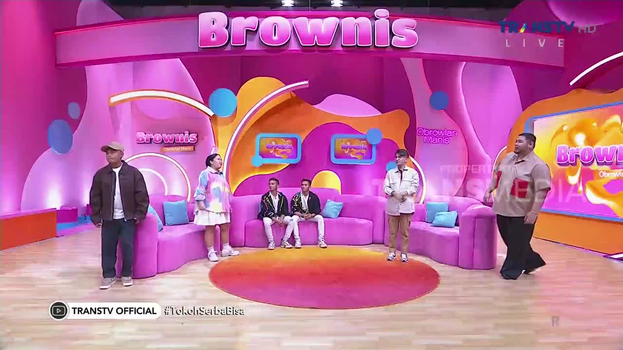 On a brightly lit set for Trans TV's "Brownis," a group of people are scattered around a pink couch. One man in a tan shirt and black pants is walking toward the right, while the others stand and face the camera.
