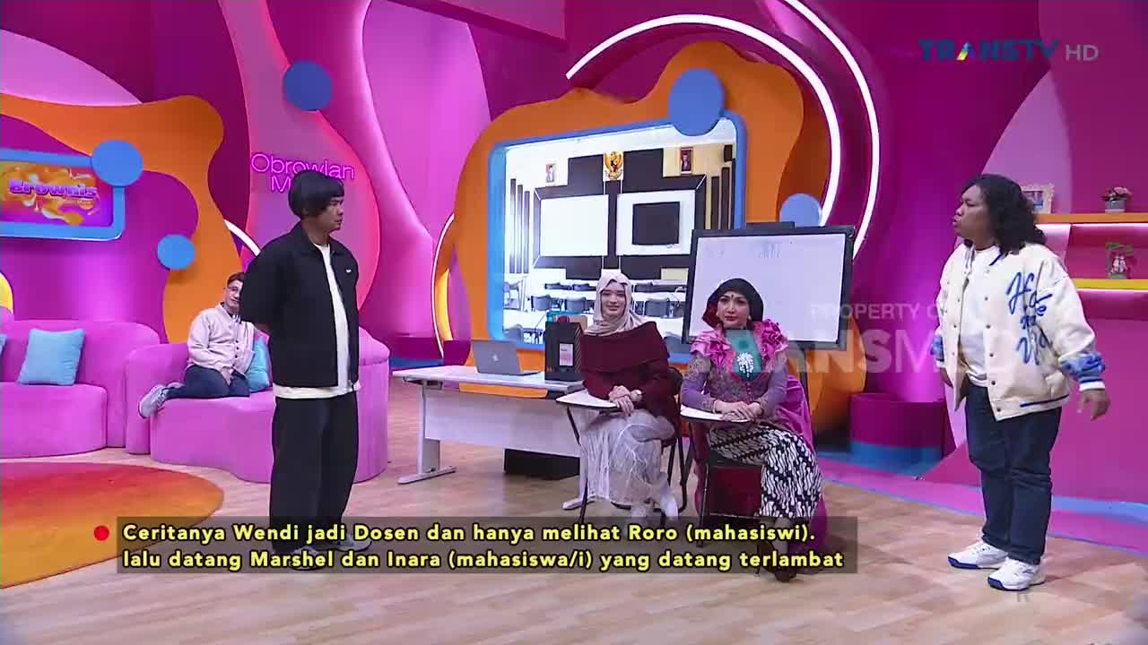 A man in a black jacket stands facing the camera, while another man in a white jacket gestures with his hands. Two women sit behind a desk, and a man lounges on a pink couch in the background.
