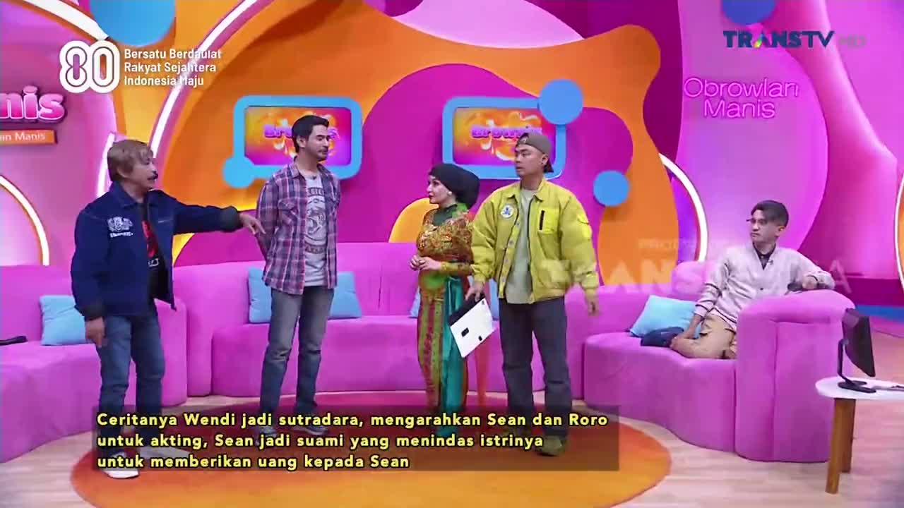 On the set of Trans TV's "Obrowlan Manis," a man in a blue jacket gestures toward two other men, while a woman in a traditional dress stands nearby. A fourth man sits on a pink couch, looking on.
