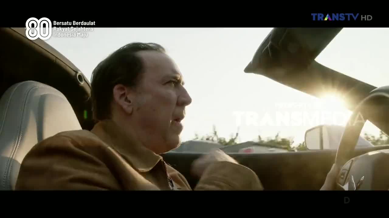 The man in the car, bathed in sunlight, looks surprised.  The Trans TV logo is visible, and the car is moving.

