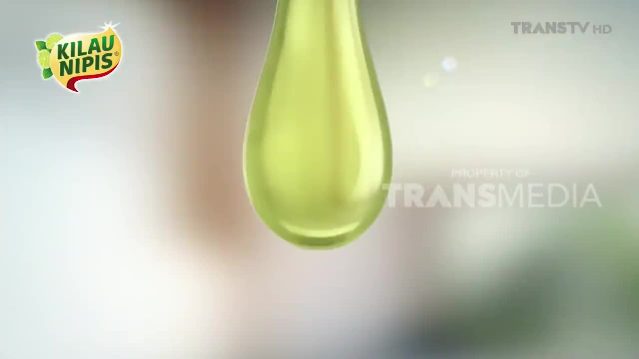 A large, translucent drop of green liquid hangs suspended, likely from a product advertisement on Trans TV Indonesia. The drop's smooth surface reflects the soft, blurred background.
