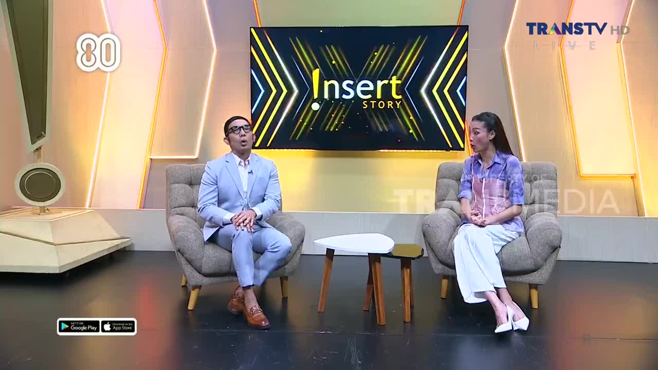 On the set of Insert Story, two people sit in armchairs facing each other. The man in the blue suit gestures with his hands as he speaks, while the woman listens intently.

