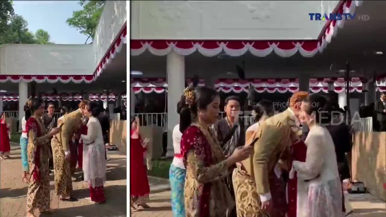 A man in a light-colored suit bows deeply as he greets women in traditional Indonesian attire. The scene, broadcast on Trans TV, appears to be a formal event, with the building decorated in red and white.

