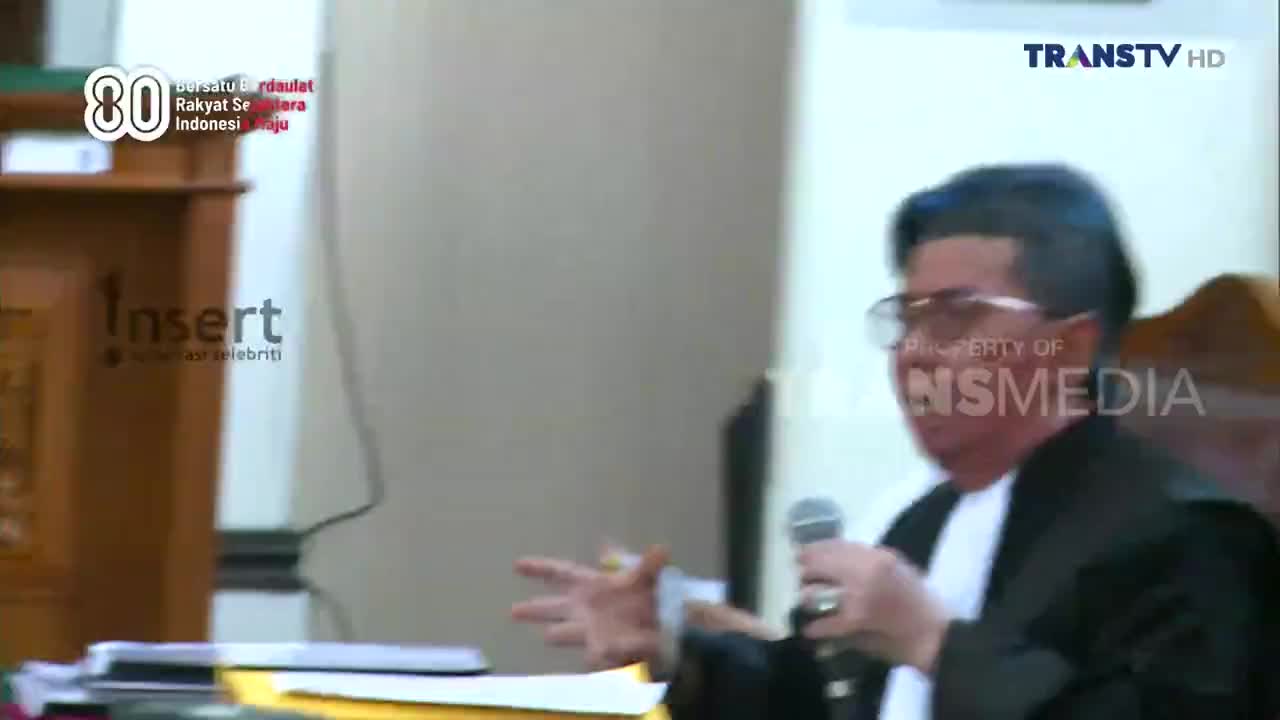 A man in a black robe gestures with his hands, holding a microphone. The scene is being broadcast on Trans TV.
