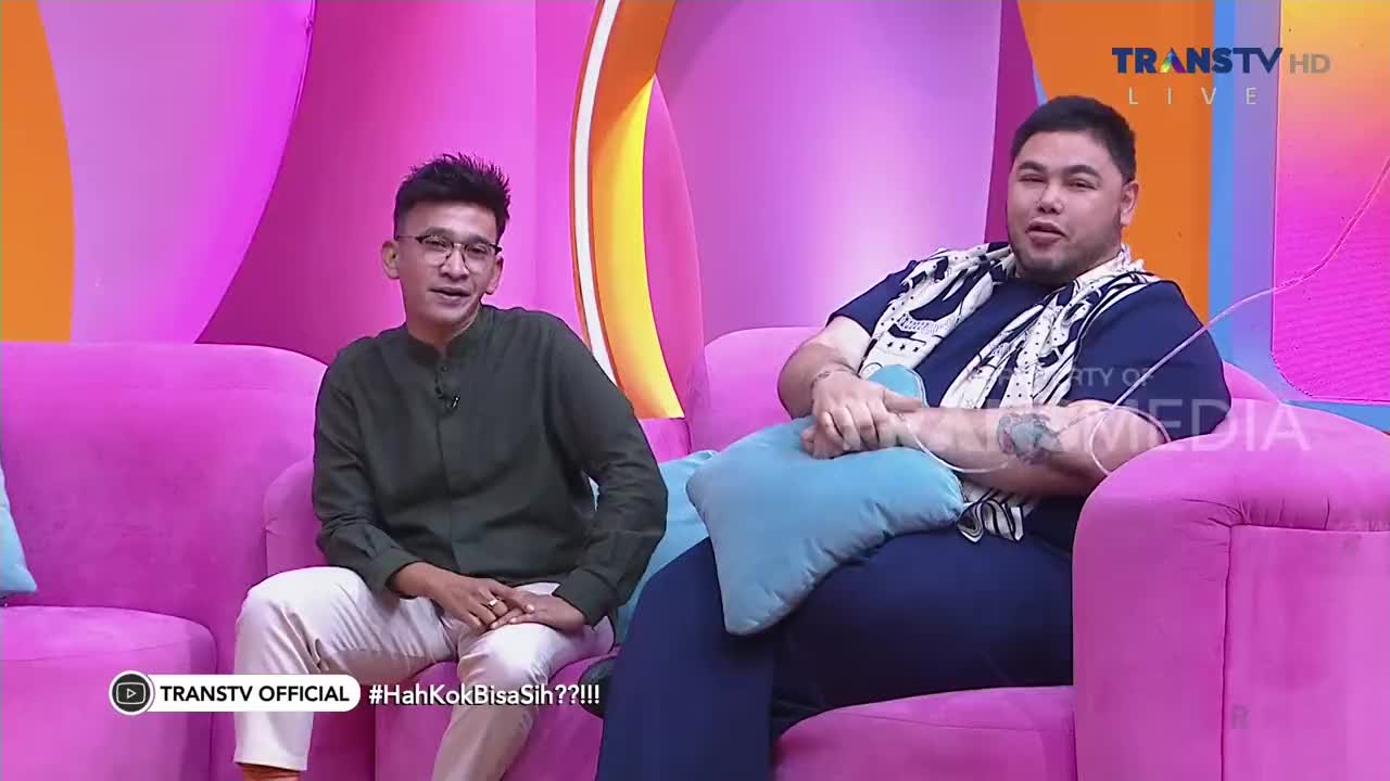 Two men are seated on a pink couch on the Trans TV set in Indonesia. The man on the right, wearing a patterned scarf, has his hands clasped in front of him.
