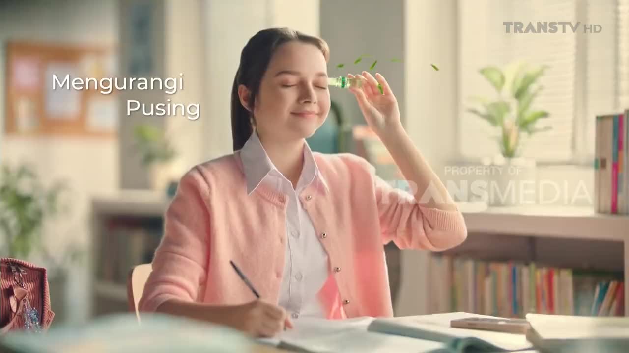 A young woman, seated at a desk, holds a small green object near her forehead, her eyes closed in a moment of relief. The scene, likely from a Trans TV commercial in Indonesia, suggests a product to alleviate headaches.
