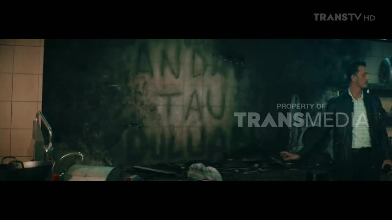 A man in a leather jacket stands in a kitchen, looking to his right, while smoke billows from a wall scrawled with Indonesian text. The Trans TV logo is visible, suggesting this scene is from a show.
