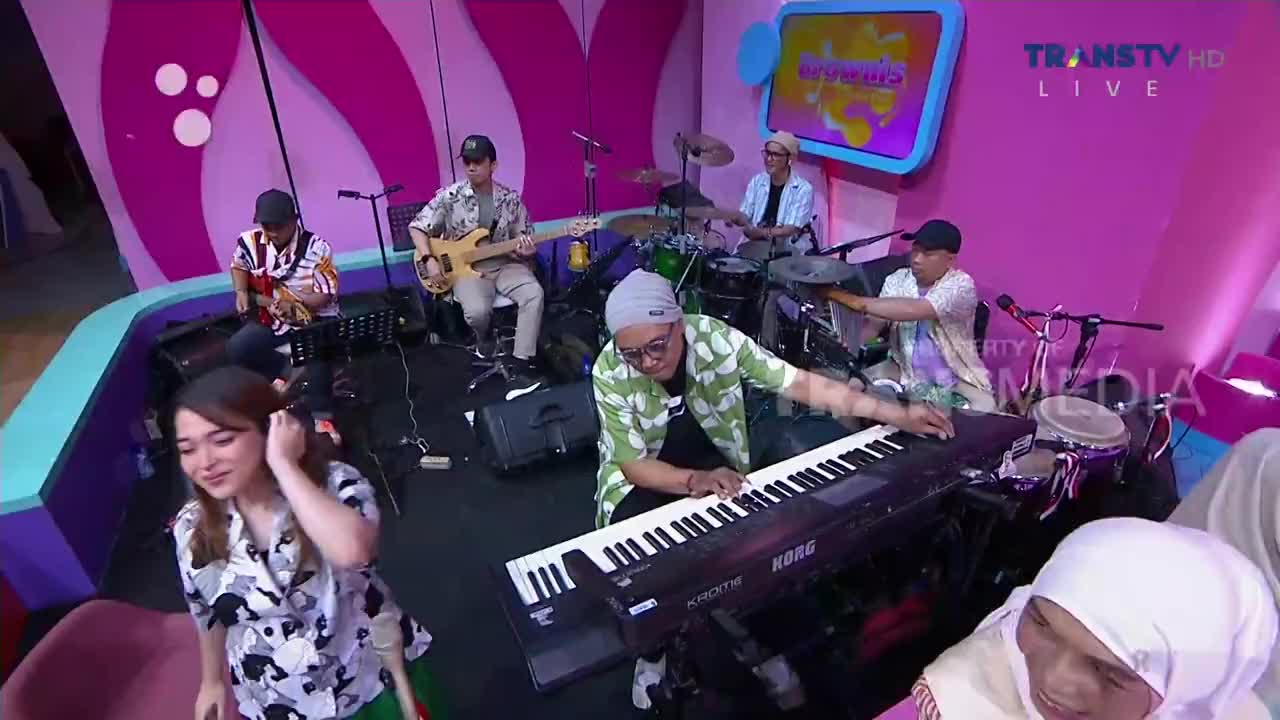 A band plays on a brightly colored set, the keyboardist hunched over his instrument. The singer smiles as she looks towards the camera, while the other musicians play their instruments.
