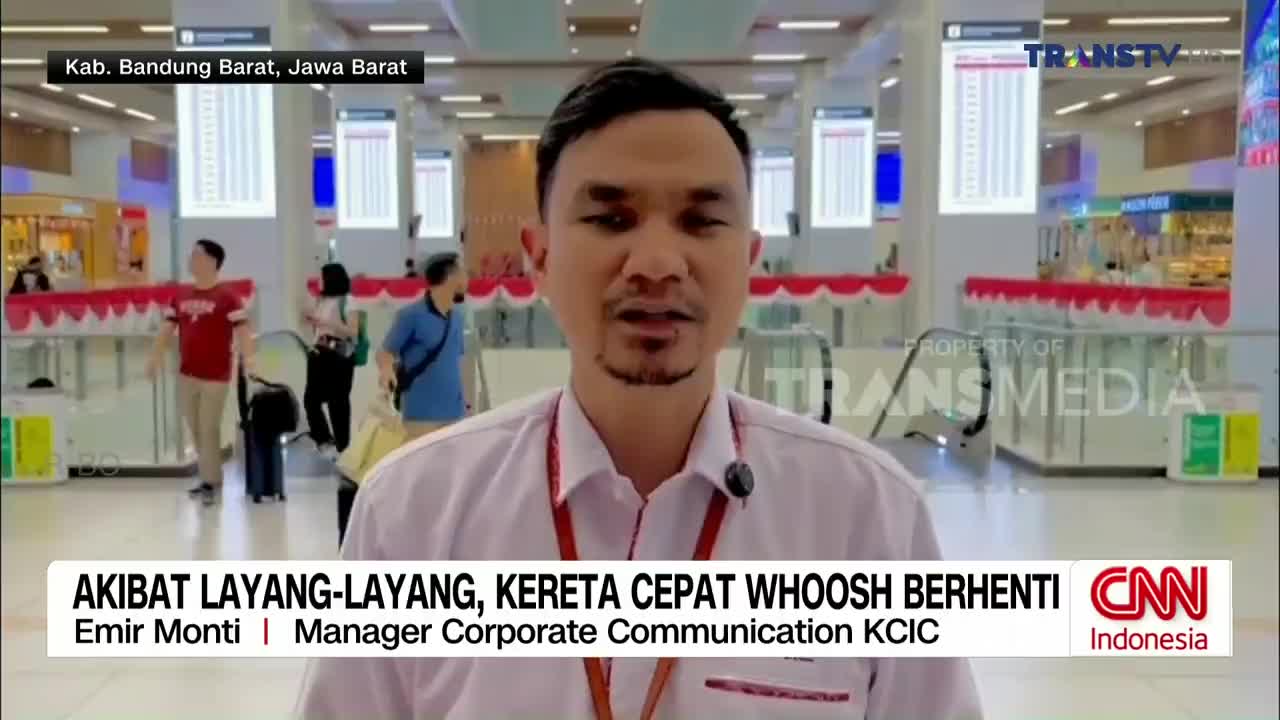 A man in a white shirt speaks directly to the camera, a Trans TV logo visible in the background. Behind him, people move through a bright, modern building, likely a train station in Indonesia.
