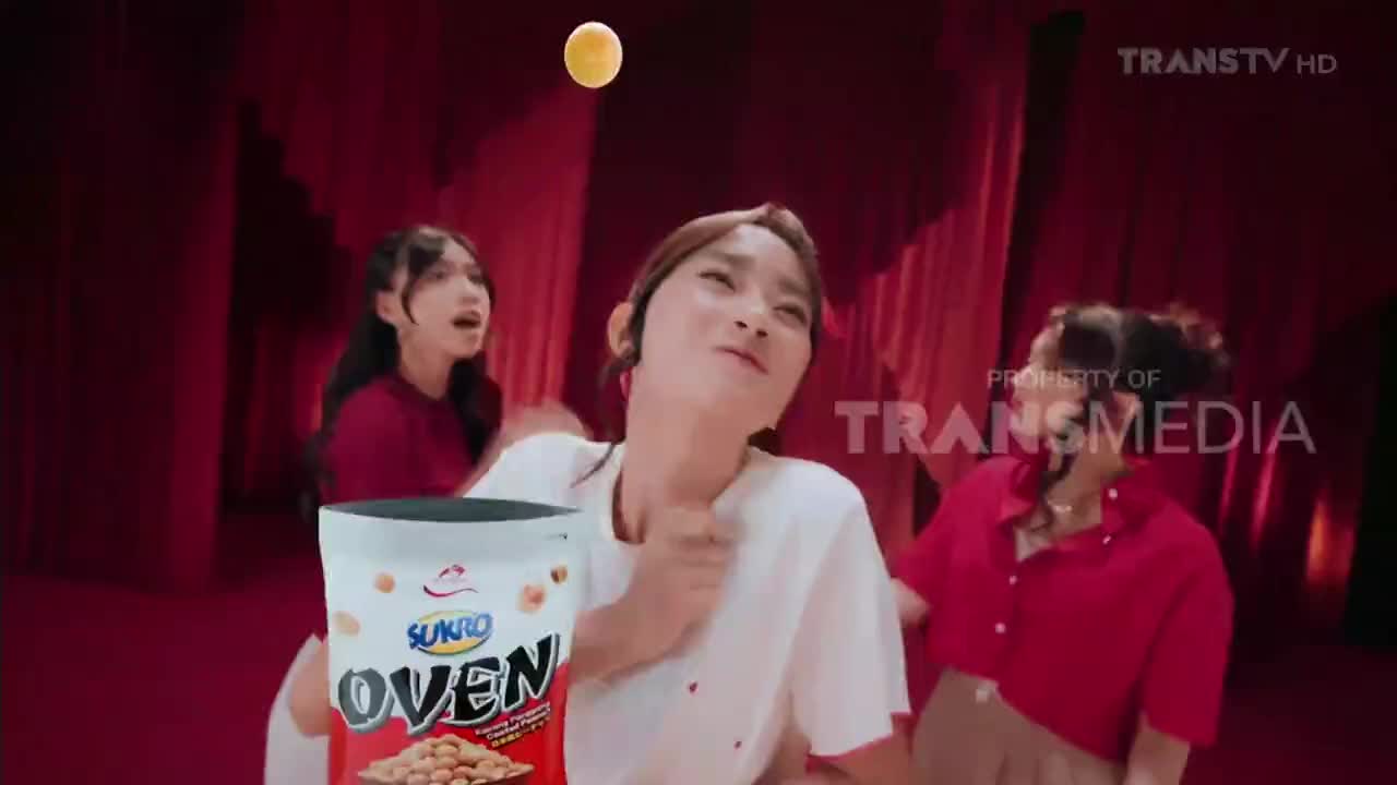 A woman in a white shirt squints upward as a yellow ball hangs in the air. Two other women in red shirts react with open mouths, all in front of a curtained backdrop, likely a commercial on Trans TV.
