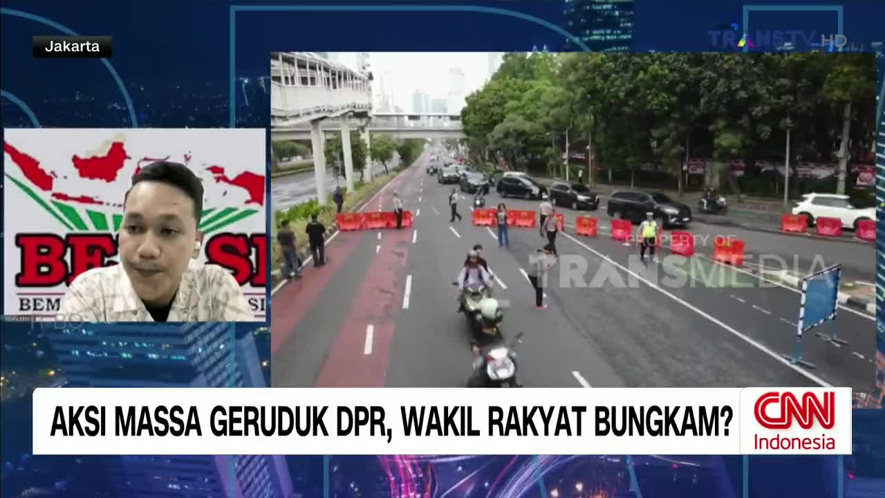 On the road, a few people walk and stand behind orange barriers. A motorcycle weaves through the stopped traffic, while a man on a screen from Trans TV discusses the situation.
