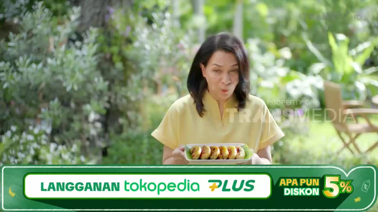 A woman in a yellow shirt holds a plate of fried bananas, blowing on them as if they're hot. The scene unfolds outdoors, likely a commercial on Trans TV in Indonesia, with a Tokopedia Plus promotion displayed.
