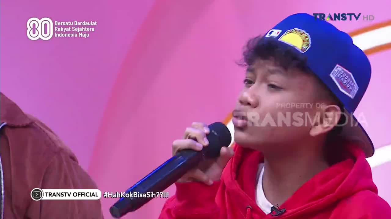 A young man in a red hoodie and a blue cap holds a microphone, singing on a Trans TV stage. He's facing forward, eyes focused, as he performs.
