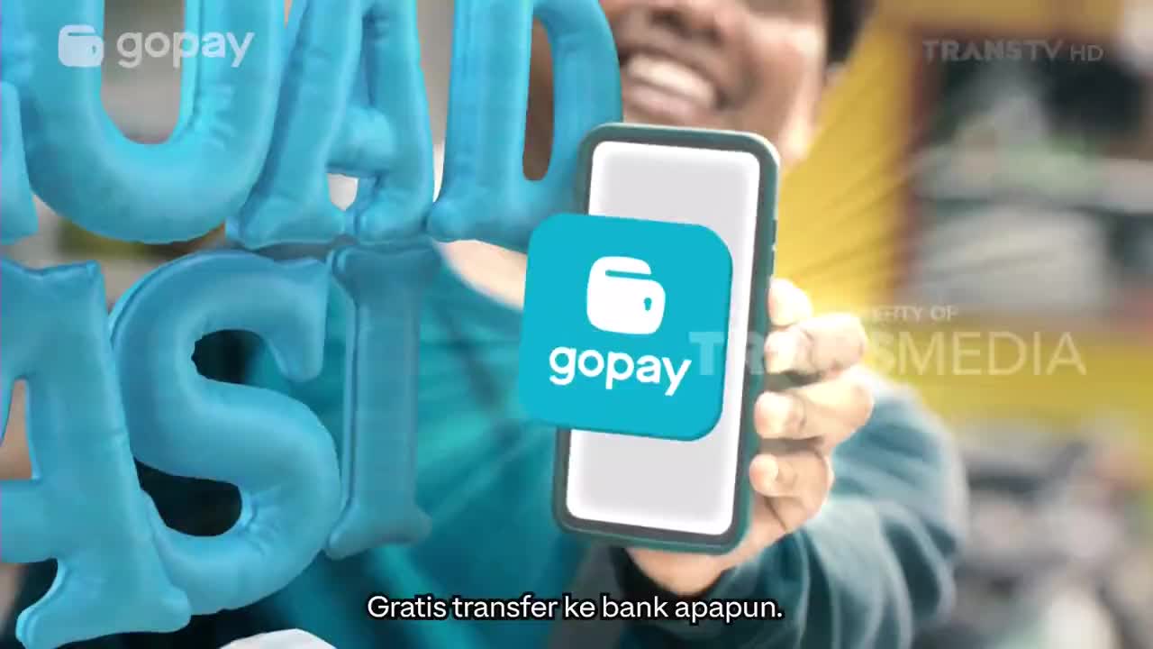 A smiling man on Trans TV in Indonesia holds a smartphone with a "gopay" logo floating in front of it. The screen displays the gopay app, and text below the phone promises free bank transfers.
