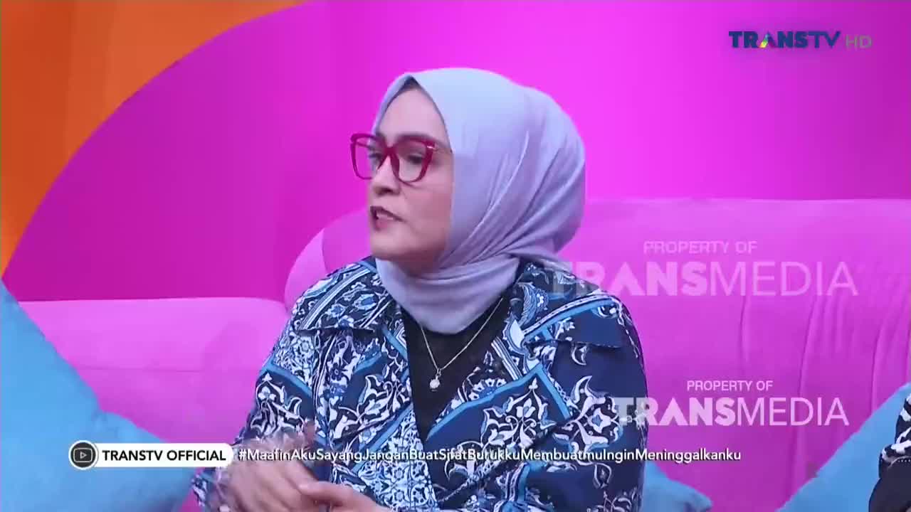 A woman in a patterned blue jacket and a light-colored hijab speaks, her red-framed glasses catching the light. The scene is set on a pink couch, likely on a Trans TV program from Indonesia.
